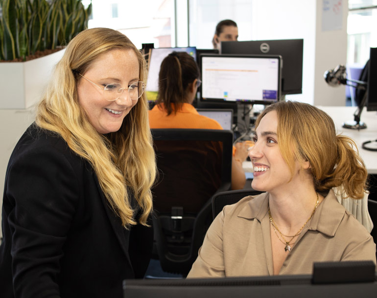 Two colleagues smiling and talking at an office desk with computer monitors in the background.