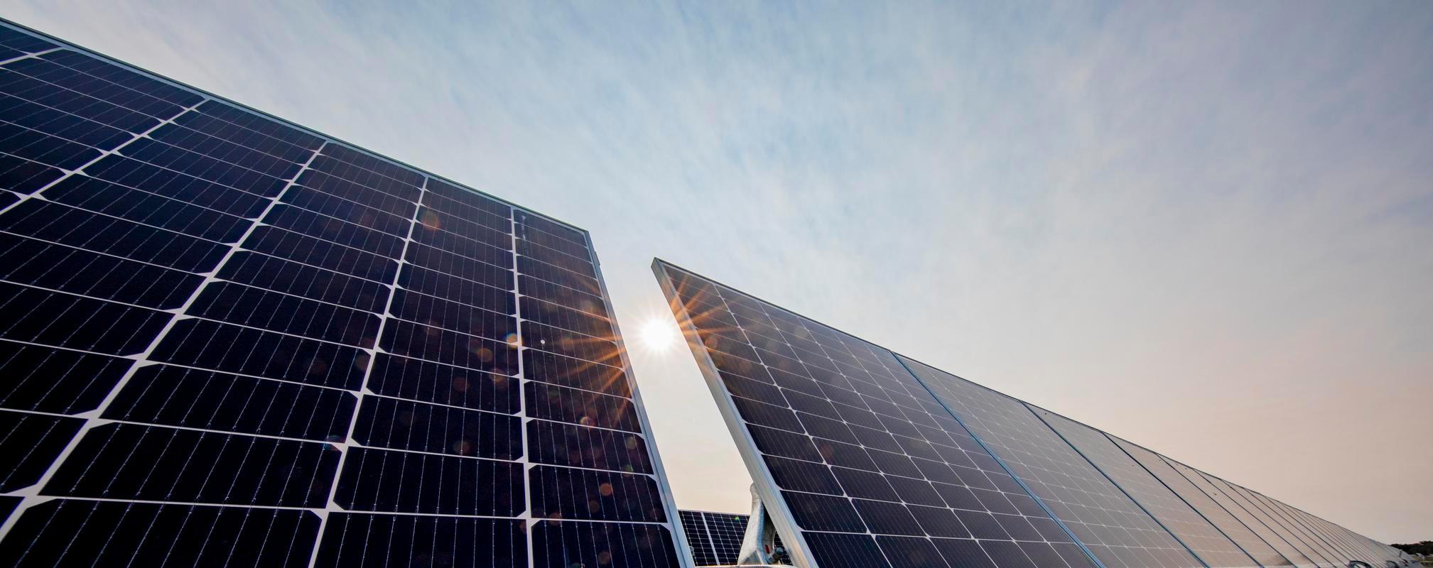 Close up, low angle shot of solar panels against a blue sky with light clouds, the sun beaming behind