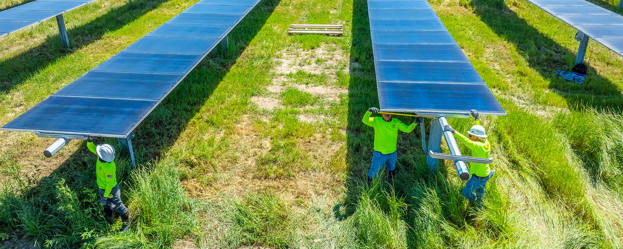 Three workers in bright green shirts and hard hats performing maintenance on solar panels installed in a grassy field.